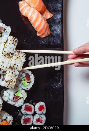 Top view of woman's hand holding sushi sticks. Sushi rolls laid out on a black plate. Vertical picture