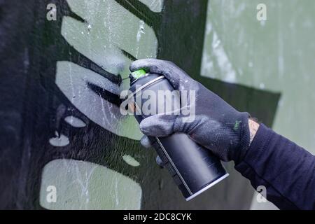 Hand of a graffiti artist in action, drawing on the wall with aerosol spray paint in a can, wearing protective gloves. Street art culture concept. Stock Photo