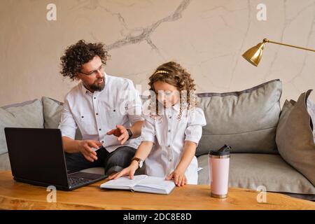 Father Works On Laptop Helps Daughter With Homework. Stay At Home During Pandemic Lockdown. High School Tutor Giving Primary School Student One To One Tuition At Desk. Stock Photo