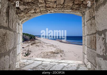 View from stone window of beach with white sand and blue sea/ocean. Beach with pine forest Stock Photo