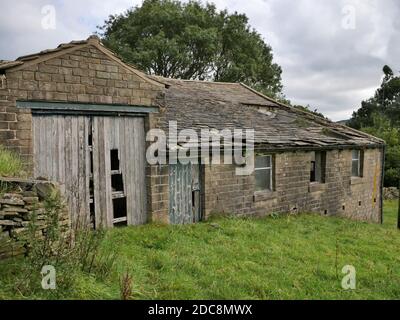 Abandoned old stone built single story barn with roof collapsed old wood doors falling apart field and trees in background Stock Photo