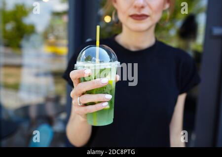 Female hand holding plastic cup with a mojito in a city cafe. Fresh cocktail drink with ice fruit and herb decoration. Stock Photo