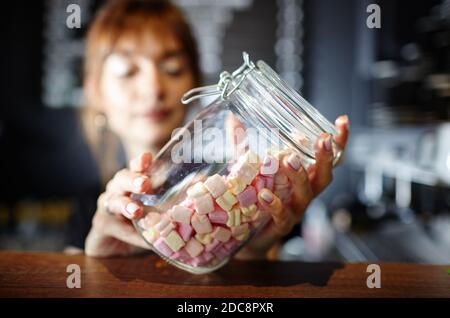 Barista holds jar with tasty mini marshmallows at bar counter in coffee shop. Blurred image, selective focus