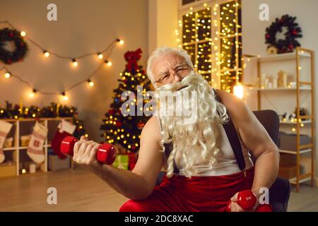 Happy Santa Claus sitting in armchair in his living-room and exercising with dumbbells Stock Photo