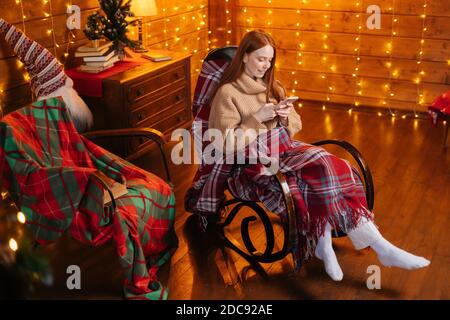 High-angle shot of relaxed young woman typing message on mobile phone sitting at cozy armchair. Stock Photo