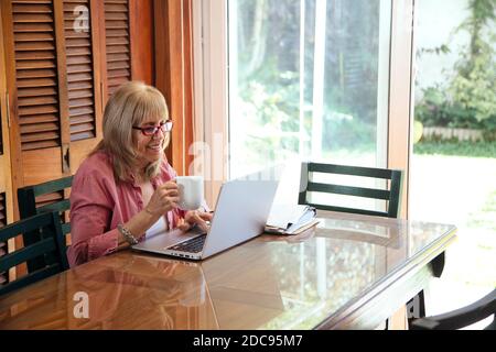 Senior woman with a laptop and coffee cup in a hand. Smiling grandmother in a wooden table with a silver computer. Stock Photo