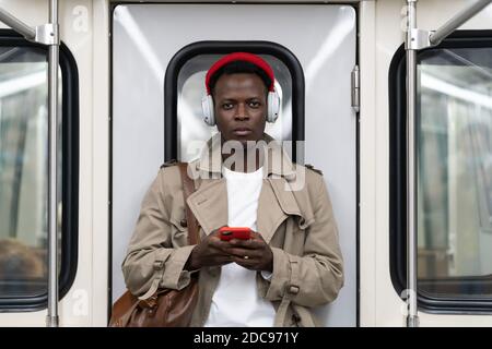 Afro-American millennial man in red hat, trench coat stand in subway train, using mobile phone, listens to music with wireless headphones in public tr Stock Photo