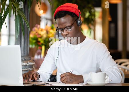 Focused millennial Black man in headphones wear red hat, watching educational webinar on laptop, remotely online work in cafe, listening audio lesson Stock Photo