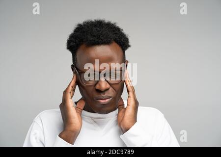 Afro man feeling headache or migraine, massaging temples, exhausted from overwork over grey background. Overwhelmed employee in glasses suffering from Stock Photo
