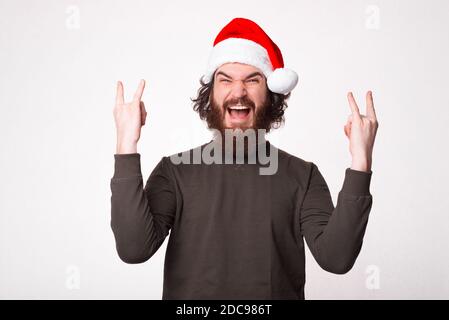 Happy bearded hipster man showing ROCK gesture over white background and wearing santa claus hat. Stock Photo