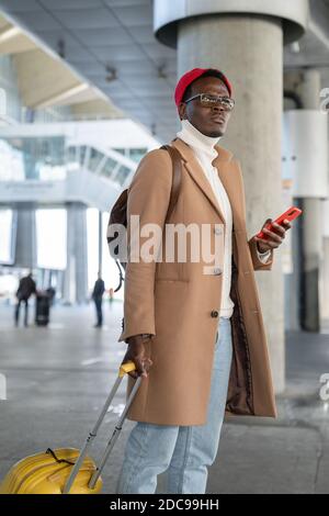 Trendy Afro-American traveler man in beige coat, red hat with yellow suitcase holding mobile phone, waiting public transportation in airport or callin Stock Photo