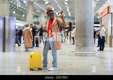 Afro-American traveler millennial man with yellow suitcase stands in airport terminal after arrival, holding mobile phone and paper cup of coffee, rai Stock Photo