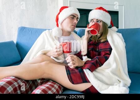 Portrait of young happy caucasian couple celebrating Christmas at home relaxing with cups of tea on a sofa wearing santa hats and pajamas Stock Photo