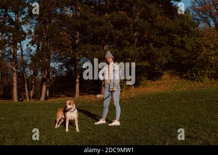 Full length of a beautiful smiling teenage girl with a hat and denim jacket posing while holding the leash of her dog breed American Staffordshire Stock Photo