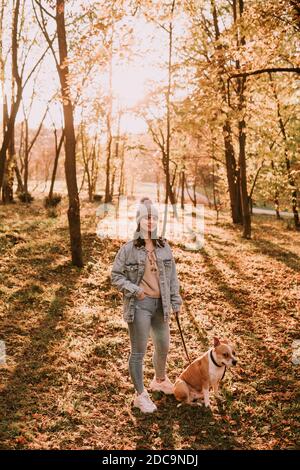 Full length of a young cute teenage girl standing in the park and holding her brown dog breed American Staffordshire Terrier. Autumn day in the woods Stock Photo