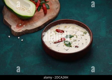 Fresh green papaya splits  arranged on a wooden piece with dry red chilli and curry leaves beside it and a wooden bowl full of raitha or pachadi made Stock Photo