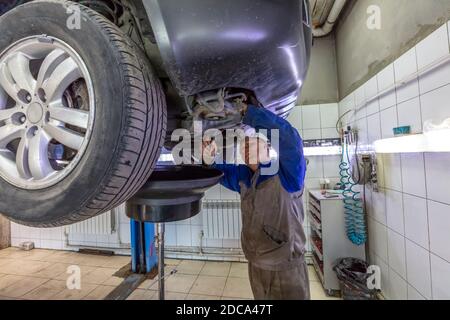 Car mechanic changes oil in a workshop. Mechanic standing under the car and draining oil into a special cannister via funnel. Stock Photo
