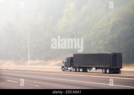 Black stylish industrial grade big rig semi truck with covered dry van semi trailer transporting commercial cargo driving on the unhealthy road in the Stock Photo