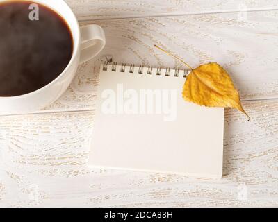 Bright yellow autumn leaf on the open spring notepad near large mug of hot steaming coffee over white wood table. Empty page for copy space. Top view. Stock Photo