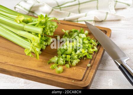 Cutting bunch of fresh green celery stalks into small pieces with a sharp knife on a brown wood cutting board over kitchen table. Natural antioxidant. Stock Photo