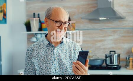 Elderly aged man waving during a video conference with family in kitchen. Elderly person using internet online chat technology video webcam making a video call connection camera communication conference call Stock Photo