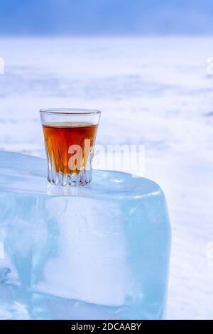 Whiskey in a glass stands on an ice block. Orange alcoholic drink in a glass. The ice melted in the sun. Light blurred background with snow. Stock Photo