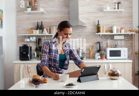 Portrait of young woman using tablet in the morning sitting at the table in the kitchen drinking tea. Working from home using device with internet technology, typing, on gadget during breakfast. Stock Photo