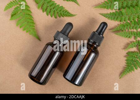 Two brown glass bottles with serum, essential oil or other cosmetic product and green fern leaves on craft beige background. Natural Organic Spa Cosme