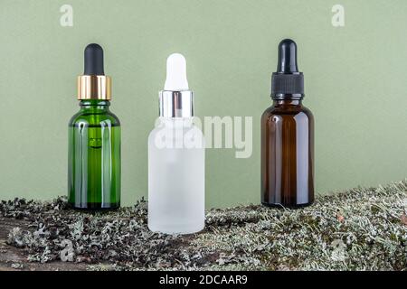 Three brown, green, white glass bottles with serum, essential oil or other cosmetic product on tree bark covered with moss against green background. N Stock Photo