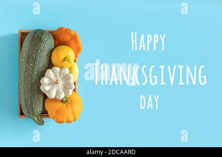 Happy Thanksgiving day text and harvest colored different vegetables gourds pumpkin, zucchini, squash in a wooden box on blue background. Top view Fla Stock Photo