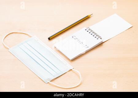 A face mask, a spiral notepad with "Covid-19" written on it and a pen are arranged on a wooden table. To-do list concept during the Covid-19 Outbreak. Stock Photo