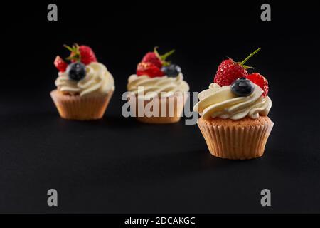 Three fresh tasty cupcakes in row isolated on black background. Closeup front view of sweet delicious dessert with white creamy topping, strawberry, blueberyy and rasberry slices. Stock Photo
