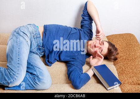 Sleepy Young Man Yawning on the Sofa with a Book Stock Photo