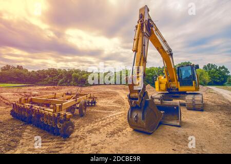 Yellow construction backhoe and disk plow heavy equipment sit in open field after the ground was modified and leveled at construction site Stock Photo