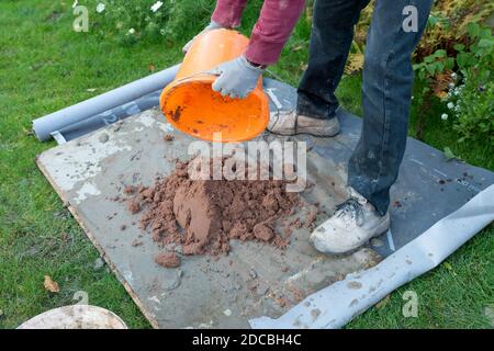close up of worker making mortar with sand and cement.  Builder with orange bucket of water tipping onto dry sand outside Stock Photo