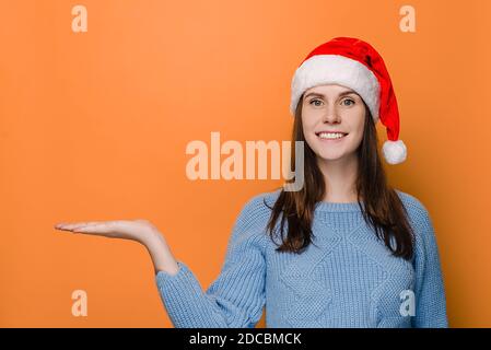 Cheerful woman with charming smile keeps palm raised against orange copy space, looks aside, wears Christmas hat and winter sweater, demonstrates some Stock Photo