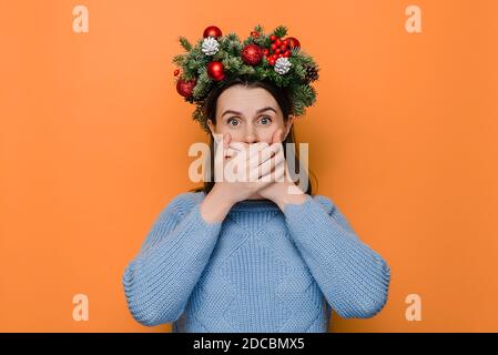 Scared emotional young woman in Christmas wreath stares at camera and covers mouth with hands cannot believe her eyes stands anxious indoor Stock Photo