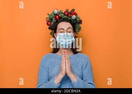 Studio portrait of young female in medical face mask prays for about the end of the coronavirus pandemic, keeps palms pressed together, eyes closed Stock Photo