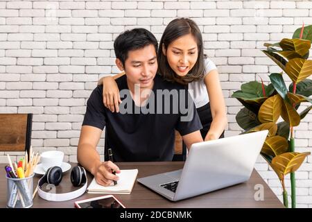 Asian woman wife motivate advice man husband freelancer working at home, sitting at desk dining table in living room Stock Photo