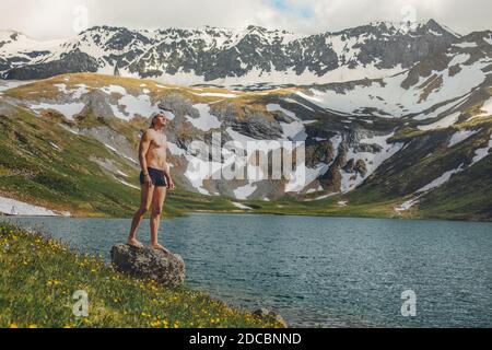 Young male tourist in a bathing suit standing up on the shore of a pure mountain lake and enjoys nature against the backdrop of snow-capped mountains. Stock Photo