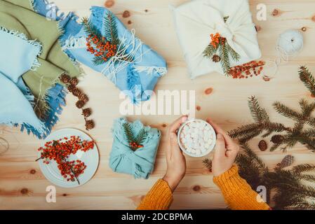 Female hands holding cup of cococa with marshmallow and just packed gift boxes wrapped in fabric and decorated with spruce branch, berries lying on Stock Photo