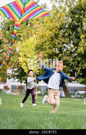Smiling boy holding flying kite while running near asian friend in park Stock Photo