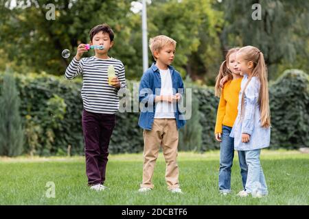 Asian boy blowing soap bubbles while standing near smiling friends on grassy lawn Stock Photo