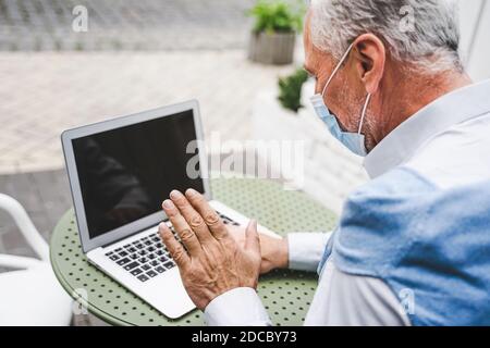 Cheerful senior man portrait with face mask during covid pandemic - Mature adult using his computer laptop in a bar coffeehouse, concepts about lifest Stock Photo