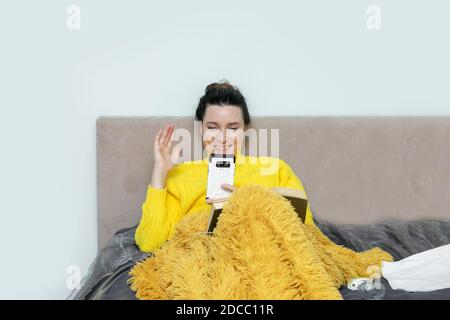 Sick cute woman on bed in sweater, under blanket in bedroom happily communicates on video call via smartphone. Self-isolation during pandemic. Concept Stock Photo