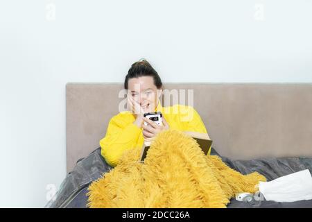 Sick cute woman on bed in sweater, under blanket in bedroom happily communicates on video call via smartphone. Self-isolation in pandemic. Concept of Stock Photo