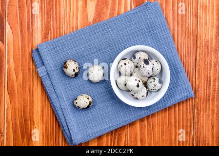 Quail eggs in white ceramic bowl on blue kitchen cloth. Top view. Detailed closeup of spotted quail eggs, rustic wooden background Stock Photo