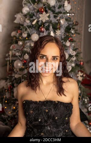 Portrait of charming brunette in black dress near Christmas tree. Young attractive woman posing at coniferous tree with decorative adornments. Concept Stock Photo