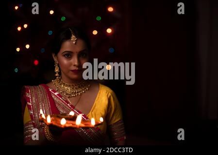 An young and beautiful Indian Bengali woman in Indian traditional dress is holding a Diwali diya/lamp in her hand in front of colorful bokeh lights. I Stock Photo