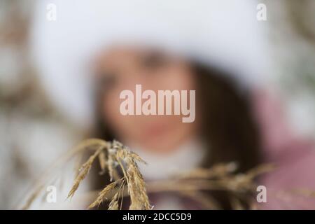 Portrait of a girl in winter clothes. In the foreground is a dried blade of grass. The girl is out of focus. Stock Photo